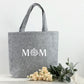 Eine elegante Shoppertasche aus Filz, personalisiert für Mütter mit Namen oder besonderem Text. Die Tasche ist ideal als Geschenkidee für Mama, Geburtstagsgeschenk, Muttertagsgeschenk oder Weihnachtsgeschenk. Das Bild zeigt die Tasche in einer gemütlichen, hellen Szene mit Blumen und Accessoires, die die stilvolle und praktische Nutzung der Tasche unterstreichen.