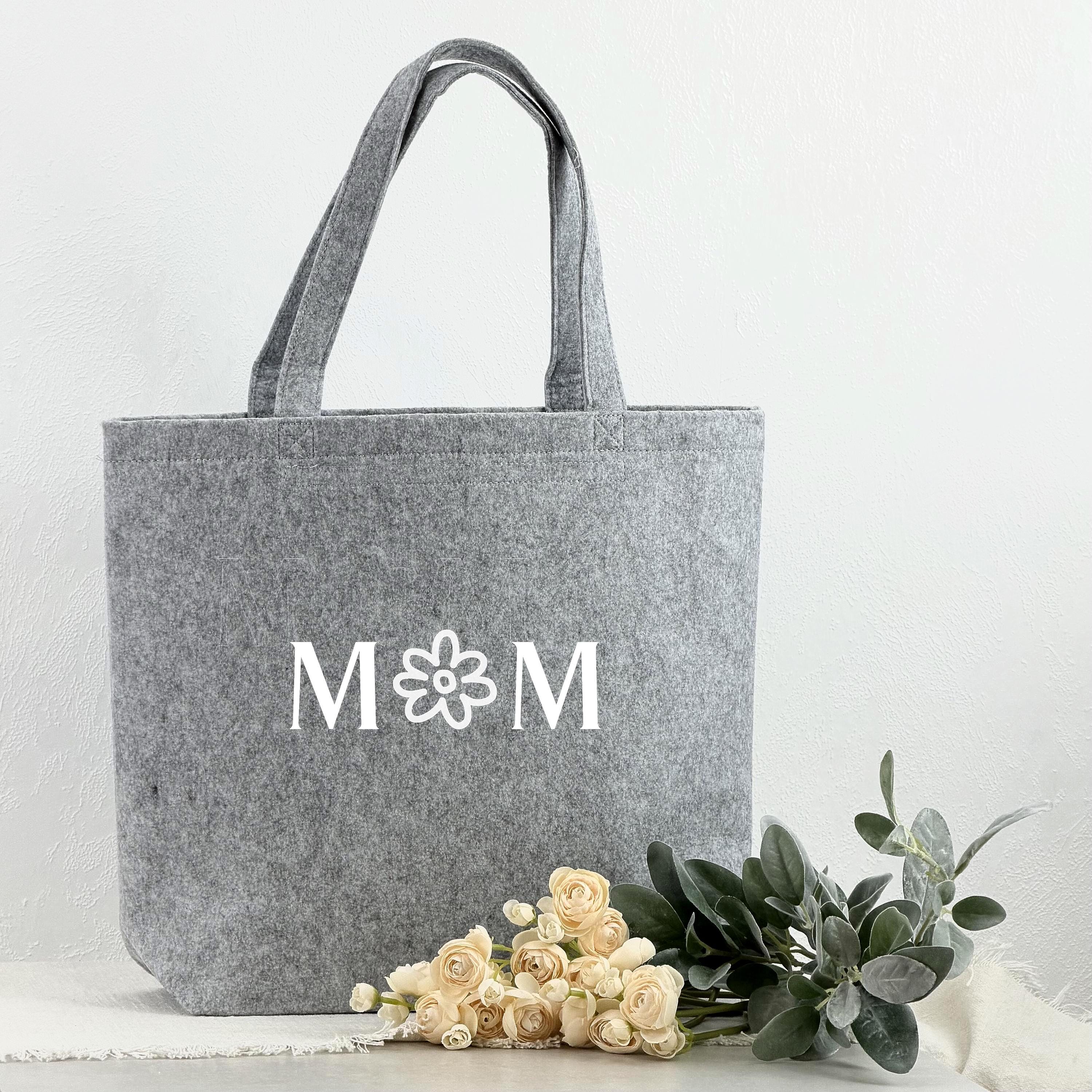 Eine elegante Shoppertasche aus Filz, personalisiert für Mütter mit Namen oder besonderem Text. Die Tasche ist ideal als Geschenkidee für Mama, Geburtstagsgeschenk, Muttertagsgeschenk oder Weihnachtsgeschenk. Das Bild zeigt die Tasche in einer gemütlichen, hellen Szene mit Blumen und Accessoires, die die stilvolle und praktische Nutzung der Tasche unterstreichen.