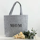 Personalisierte Shoppertasche aus Filz, liebevoll gestaltet für Mama als Geburtstags- oder Muttertagsgeschenk. Das Foto zeigt die Tasche auf einem Holztisch mit weichen Lichtakzenten und Dekorationen wie Blumen oder Kerzen. Die Gravur oder Stickerei macht sie zu einem emotionalen Geschenk, das praktische Funktionalität und stilvolles Design perfekt kombiniert.