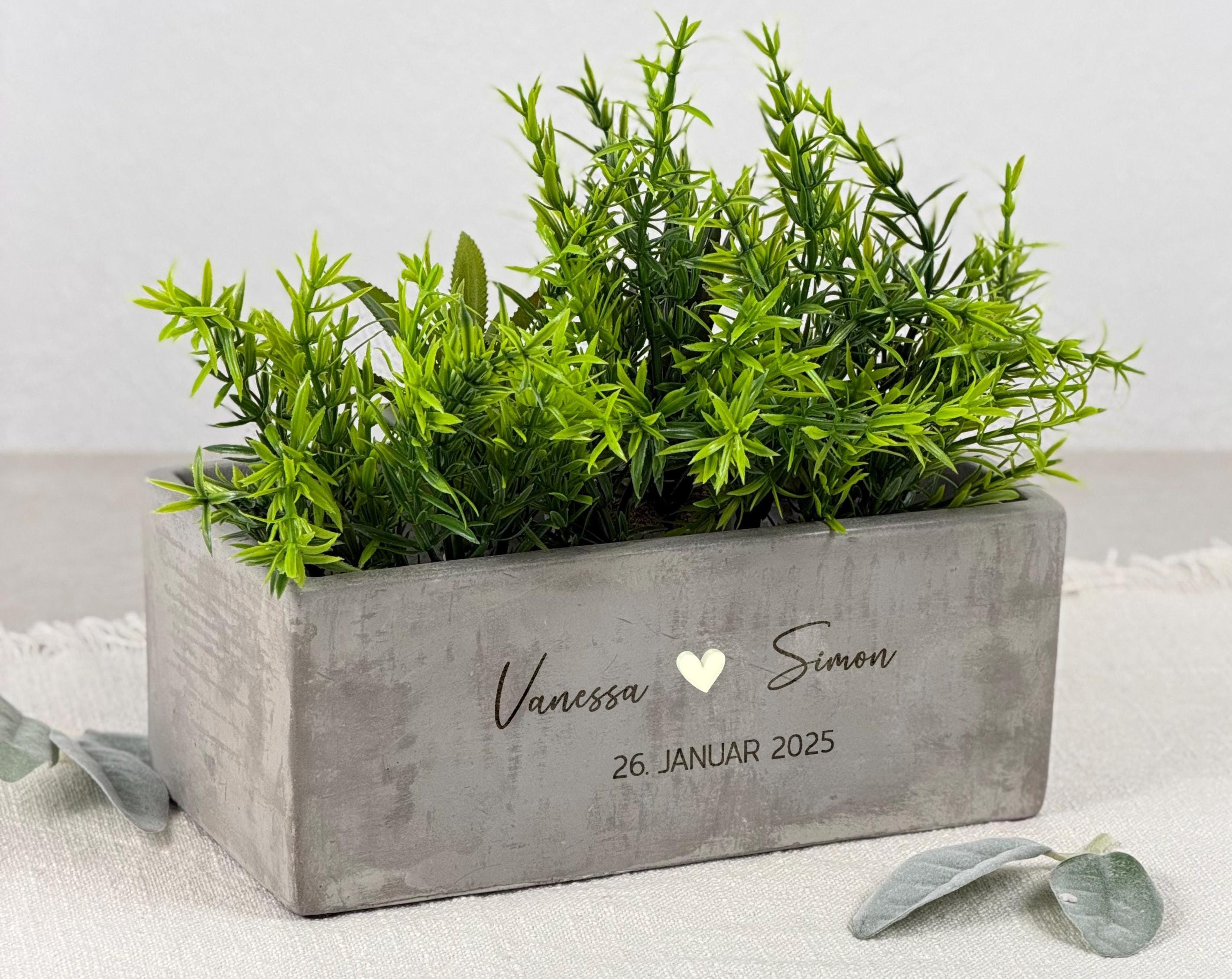 Personalisierter Pflanztopf für Paare, darauf fein gravierte Namen und ein romantischer Spruch. Der Blumentopf steht auf einer hellen Holzfläche und wird von einer kleinen grünen Pflanze ergänzt. Ein ideales Hochzeitsgeschenk, Einzugsgeschenk oder Geschenk zur Verlobung. Das moderne, minimalistische Design macht ihn zu einer zeitlosen Deko-Idee für Wohnzimmer, Küche oder Balkon.