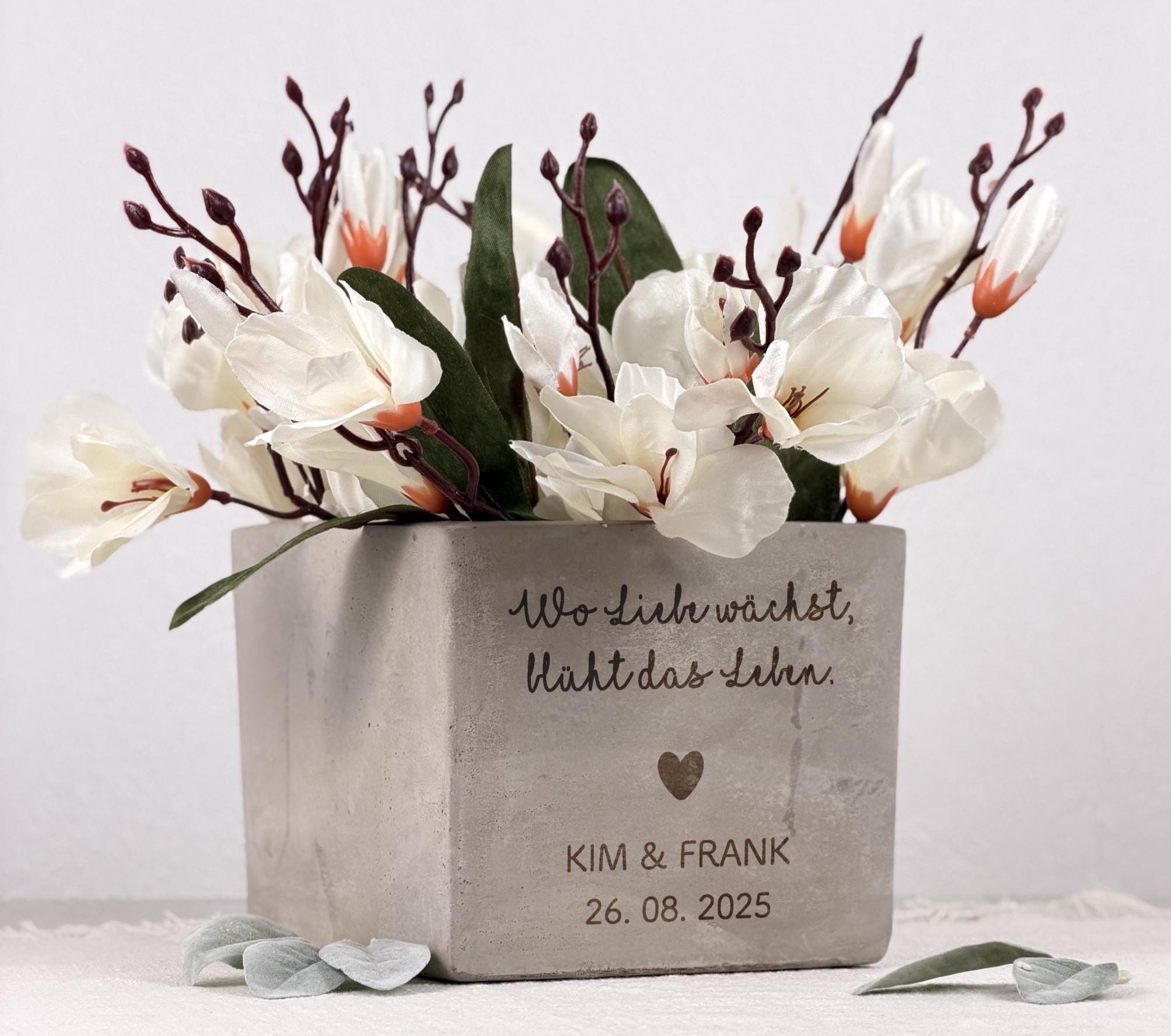 Ein personalisierter Blumentopf mit graviertem Namen des Brautpaars, gestaltet als besonderes Hochzeitsgeschenk. Der Topf zeigt eine elegante Oberfläche, einen warmen Naturton und eine liebevolle Gravur mit Herzen und Datum. Eine kleine Pflanze sitzt dekorativ im Inneren. Ideal als Geldgeschenk-Idee oder Einweihungsgeschenk, perfekt für Paare, die ein stilvolles und persönliches Dekostück für ihr neues Zuhause suchen.