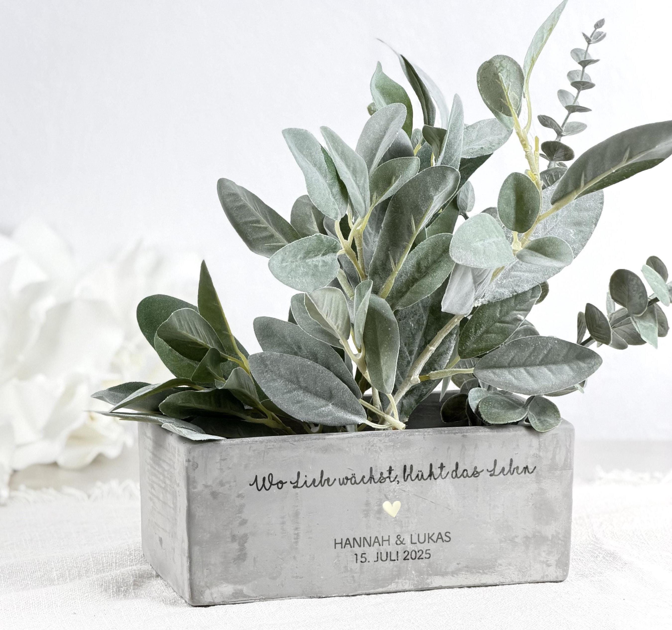 Dekorativer Blumentopf mit personalisiertem Namen für Paare, als liebevolles Geschenk zur Hochzeit. Der Topf hat eine moderne Form und eine feine Gravur mit Herzen. Neben einer kleinen Pflanze wirkt er warm und einladend. Perfekt als Paargeschenk, Valentinstag-Geschenk oder Geschenk zur Verlobung. Hochwertiges Material, ideal für romantische Deko im Wohnzimmer oder Schlafzimmer.
