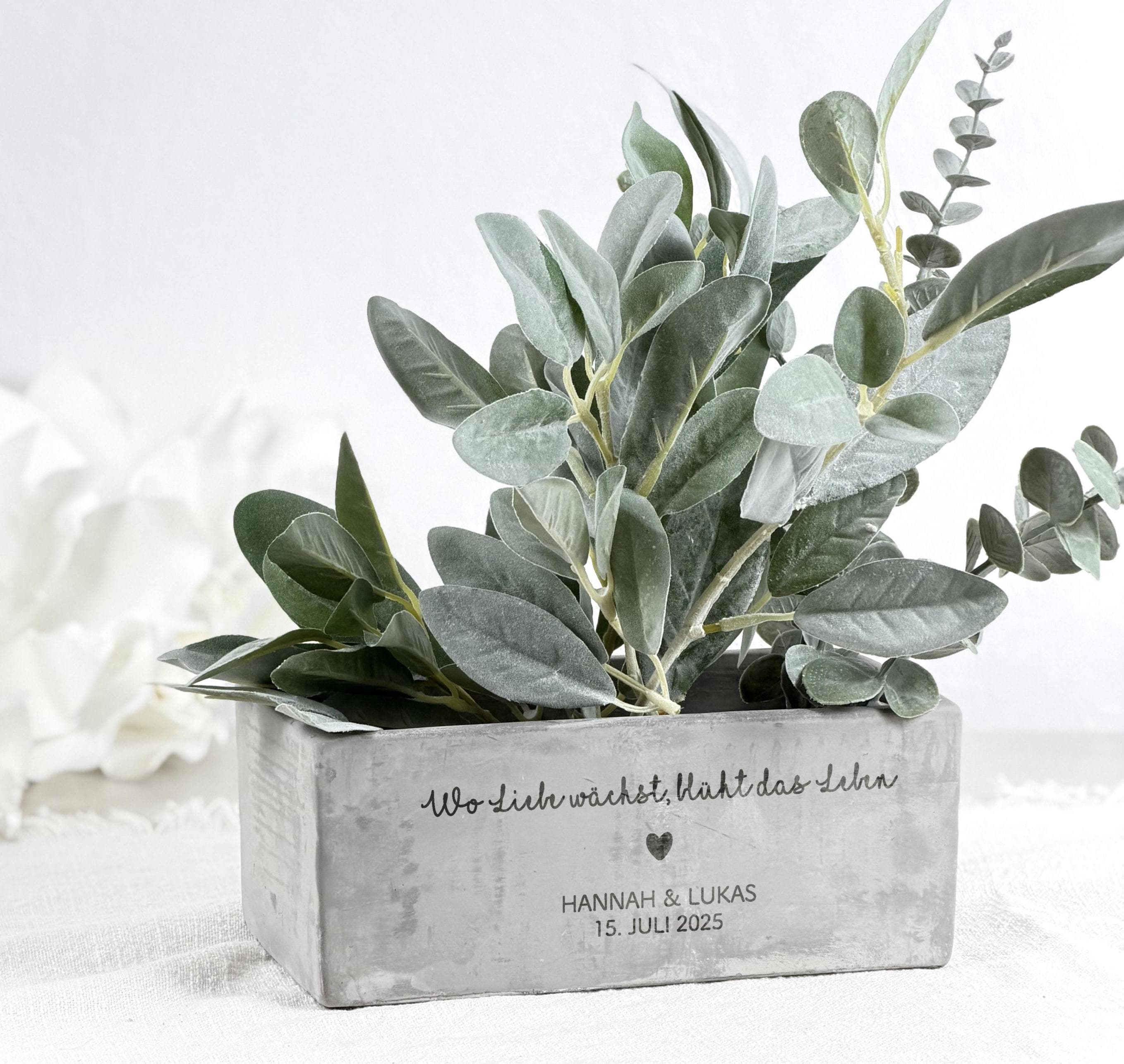 Personalisierter Blumentopf mit Gravur, dargestellt als Geschenkidee zur Hochzeit oder zum Einzug. Der Topf trägt einen individuellen Namen und ein Datum und enthält eine kleine dekorative Pflanze. Die warme Farbe des Topfes und die klare Gravur verleihen ihm ein elegantes, zeitloses Erscheinungsbild. Perfekt für Brautpaare, frisch Verlobte oder Paare, die ein neues Zuhause feiern.