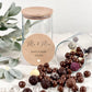 Ein transparentes Vorratsglas mit personalisierter Gravur, perfekt für ein Paar. Die Gravur enthält Namen, ein kleines Herzmotiv sowie ein Datum. Das Glas steht im Mittelpunkt des Bildes, mit weichem Hintergrundlicht und pastellfarbenen Accessoires. Dieses Foto vermittelt die Eleganz und Emotionalität eines Geschenks, das ideal für Valentinstag, Hochzeiten, Geldgeschenke oder Jubiläen geeignet ist. Ein stilvolles und bedeutungsvolles Präsent für Partner.
