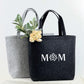Ein personalisiertes Mama Geschenk: eine hochwertige Filztasche mit individuell eingesticktem Namen oder Wunschtext. Die Tasche ist stilvoll, robust und vielseitig einsetzbar für Alltag, Shopping oder Freizeit. Perfekt als Geburtstagsgeschenk, Muttertagsgeschenk oder Weihnachtsgeschenk. Das Foto zeigt die Tasche auf einem hellen Hintergrund mit dekorativen Elementen, die Wärme, Liebe und Praktikabilität vermitteln.